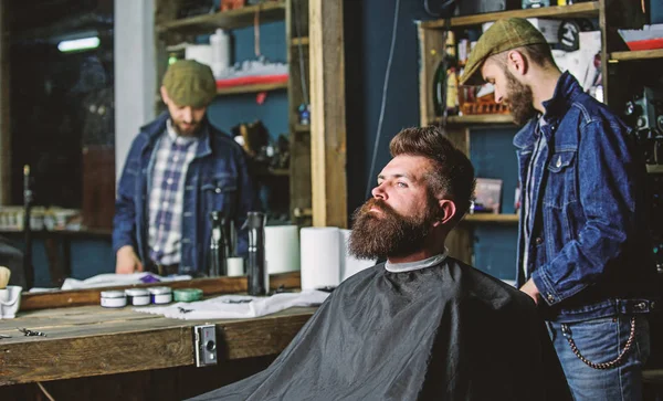Hipster client getting haircut. Man with beard covered with black cape waiting while barber changing clipper grade. Client with beard ready for trimming or grooming. Haircut process concept