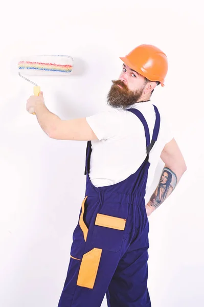 Appartement herontwerp en reparatie. Weer aannemer in oranje helm staande met verfroller in zijn hand. Geïsoleerde bebaarde man met tatoeage op zijn arm op witte achtergrond — Stockfoto