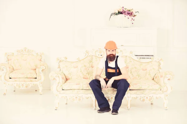 Loader sit on sofa, having rest. Break and rest concept. Man with beard, worker in overalls and helmet sitting on couch tired, white background. Courier relaxing while moving furniture, relocation.