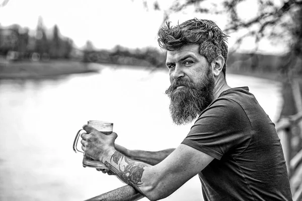 Side view bearded man enjoying big mug of beer on warm evening. Brutal man with messy hairstyle and tattooed arm having a drink in bar on the river font. Friday night after work — Stock Photo, Image