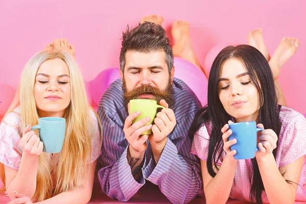 Lovers drinking coffee in bed. Threesome relax in morning with coffee. Stormy night concept. Man and women in domestic clothes, pajamas. Man and women, friends on sleepy faces lay, pink background