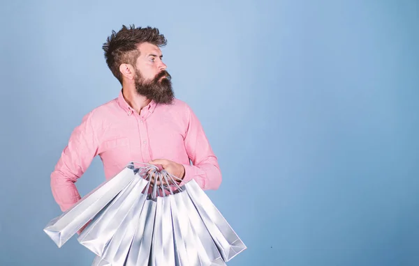 Sale and discount concept. Hipster on serious face carries shopping bags. Man with beard and mustache works as shop assistant, light blue background. Guy shopping on sales season with discounts