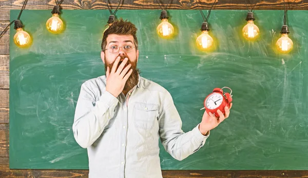 Forgot about time concept. Man with beard and mustache on shocked face in classroom. Bearded hipster holds clock, chalkboard on background, copy space. Teacher in eyeglasses holds alarm clock — Stock Photo, Image