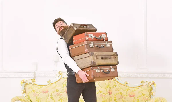 Mann mit Bart und Schnurrbart im klassischen Anzug liefert Gepäck, luxuriöses weißes Interieur. Butler und Servicekonzept. Macho elegant auf überraschtem Gesicht trägt Stapel alter Koffer — Stockfoto