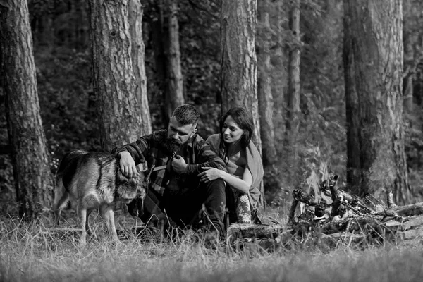 Man and woman traveling with dog at camp. Happy couple travel with dog in nature. Smiling young people sitting near bonfire and playing with pet, traveling on weekend.