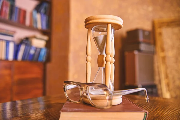 Пісок падає всередину пісочного годинника. Пісочний годинник, стара книга та окуляри на дерев'яному столі, інтер'єр на фоні. Вчені атрибути концепції. Годинник підраховує час біля старомодних окулярів — стокове фото