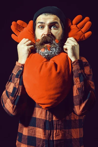 Bel homme barbu ou un gars en chapeau d'hiver et chemise à carreaux avec moustache à la mode sur le visage surpris et coeurs rouges décorés avec de la neige dans la barbe tient oreiller de valentines sur fond noir — Photo