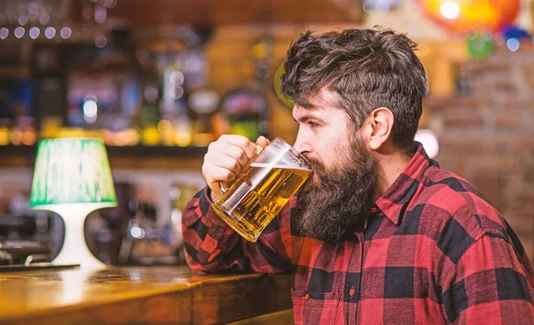 Tipo pasar el ocio en el bar, fondo desenfocado. Hipster con barba sostiene el vaso con cerveza, bebidas, disfrutar de un sabor fresco. Concepto de relajación. El hombre en la cara tranquila se sienta cerca del mostrador del bar, sacia la sed —  Fotos de Stock