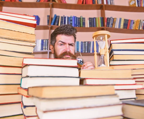 Man, scientist peeking out of books. Teacher or student with beard studying in library. Man on serious and strict face watching time is going over, bookshelves on background. Time flow concept
