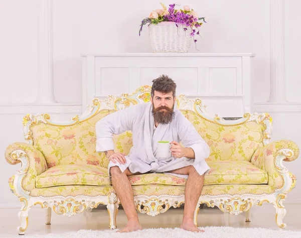 Человек с бородой и усами наслаждается утром, сидя на роскошном диване. Мужчина на сонном строгом лице в халате, пьет кофе, в роскошном отеле утром, на белом фоне. Элитный досуг — стоковое фото