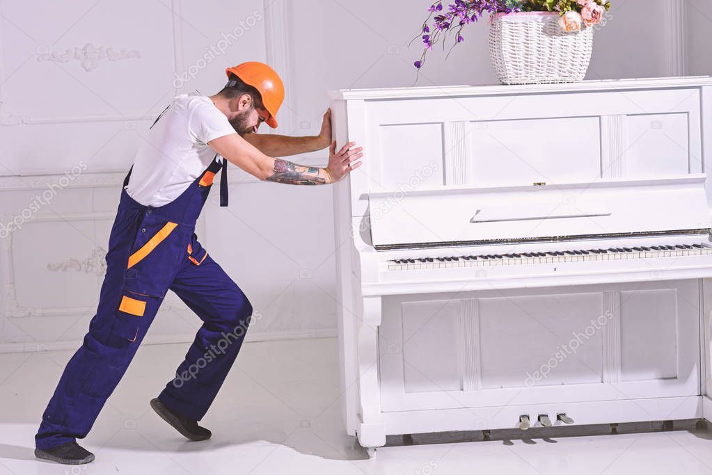 Heavy loads concept. Loader moves piano instrument. Courier delivers furniture, move out, relocation. Man with beard worker in helmet and overalls pushes, put efforts to move piano, white background