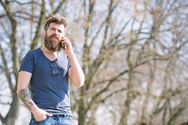 Hipster with beard speak on mobile phone outdoor. Man with beard and mustache on strict face talking, nature background, defocused. Bearded man speaking on cell phone. Communication concept