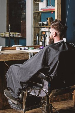 Hipster berber sakal müşterilerinden olan adam. Sakallı hippi Kuaför ve saç kesimi için bekler. Berber kavramı. Siyah pelerin ile kaplı sakallı adam ayna önünde salonların sandalyede oturur