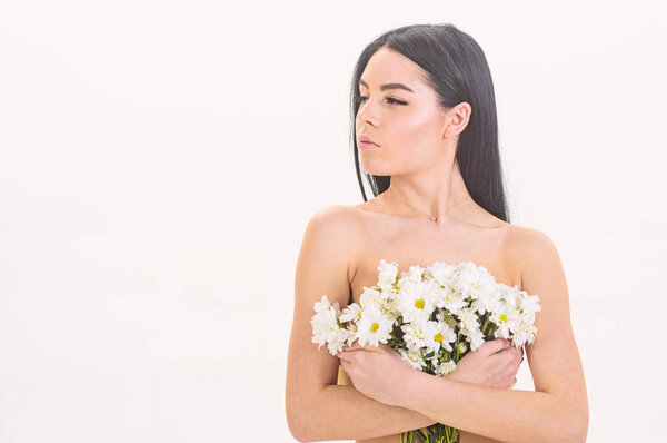 Girl on calm face stands naked and holds chamomile flowers in front of chest. Lady covers breasts with flowers, isolated on white. Skin health concept. Woman with smooth healthy skin looks attractive