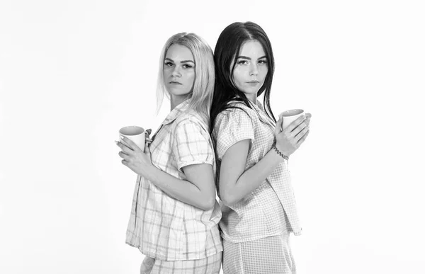 Conceito de café da manhã. Loira e morena em rostos sonolentos segura canecas com café. Irmãs ou melhores amigas de pijama ficam de costas para trás. Meninas bebendo café de manhã, fundo branco isolado — Fotografia de Stock