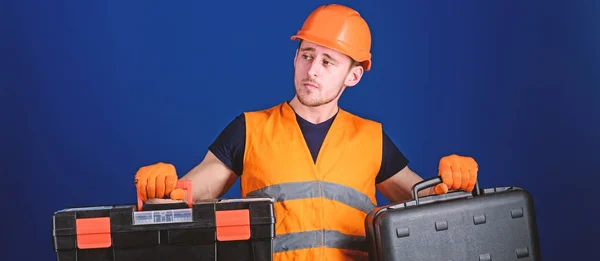 Concetto di cassetta degli attrezzi e attrezzature. Operaio, riparatore, riparatore, costruttore su viso premuroso scegliendo attrezzature per il lavoro. Uomo in casco, cappello rigido porta attrezzi e valigia con attrezzi, sfondo blu — Foto Stock