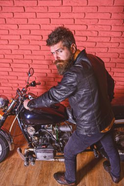 Yenilikçi, vahşi Motorcu deri ceketli sıkı yüzündeki motosiklet üzerinde oturur. Sakallı, motorcu motorlu bisiklet garaj, tuğla duvar arka plan yakınındaki deri ceketli adam. Erkeksi hobi kavramı