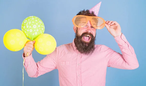 Hipster em óculos gigantes celebra o aniversário. Partido atribui conceito. Homem com barba no rosto feliz segurar balões de ar, fundo azul claro. Cara em chapéu de festa com balões de ar celebra — Fotografia de Stock