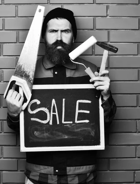 Bearded foreman holding various building tools and board, serious face — Stock Photo, Image