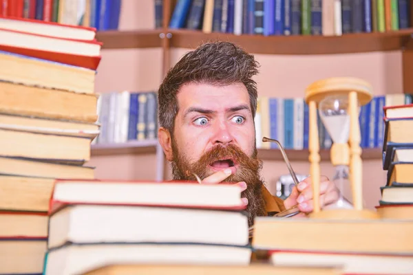 Mann, Wissenschaftler schaut auf Sanduhr. Lehrer oder Schüler mit Bart studieren in der Bibliothek. Mann mit ernstem und schockiertem Gesicht beobachtet, wie die Zeit vergeht, Bücherregale im Hintergrund. Zeitflusskonzept — Stockfoto