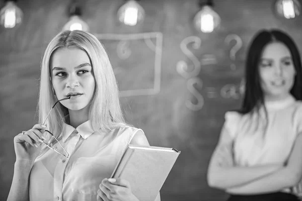 Mädchen eifersüchtig auf den Erfolg ihrer Klassenkameradin im Klassenzimmer, Kreidetafel im Hintergrund. Frau mit Buch und Brille sieht verträumt aus, während Studentin im Hintergrund eifersüchtig guckt. Neid und Konkurrenzdenken — Stockfoto