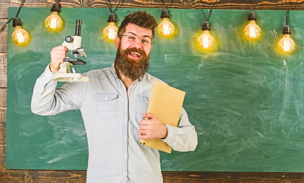 Profesor en anteojos sostiene libro y microscopio. Científico sostiene libro y microscopio, pizarra en el fondo, espacio de copia. Concepto científico. Hombre con barba y bigote en cara sonriente en clase — Foto de Stock