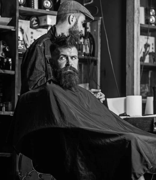 Um cliente Hipster a cortar o cabelo. Processo de corte de cabelo conceito. Homem com barba coberta com capa preta esperando enquanto o barbeiro muda de categoria clipper. Cliente com barba pronta para aparar ou arrumar — Fotografia de Stock