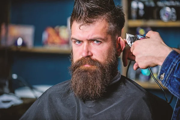 Barber works with hair clipper. Barbershop concept. Hands of barber with clipper, close up. Hipster bearded client on strict face getting hairstyle. Man with beard as client, barbershop background