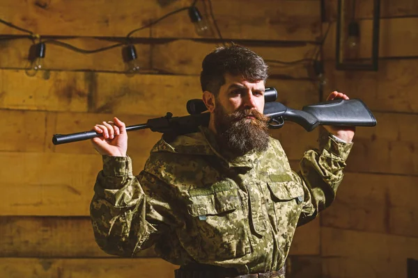 Hunter, brutal hipster on strict face with gun ready for hunting. Man, gamekeeper with beard wears camouflage clothing, carries rifle on shoulders, wooden interior background. Masculinity concept