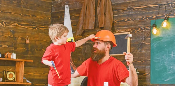 Father, parent with beard in protective helmet teaching son to use different tools in school workshop. Fatherhood concept. Boy, child cheerful holds toy saw, having fun while handcrafting with dad