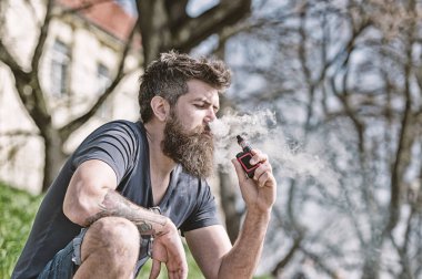 Sakallı adam vape güneşli günde sigara içiyor. Gevşeme kavramı. Uzun sakallı ve bulutlar duman görünüyor rahat olan adam. Adam sakal ve bıyık sakin yüzündeki defocused arka plan, dallarda