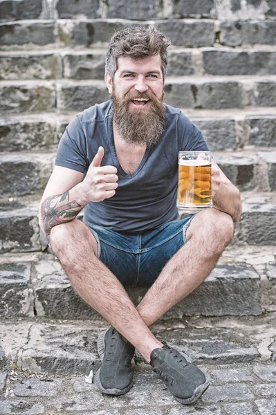 Hombre con barba y bigote en la cara feliz, fondo pedregoso, desenfocado. Hombre barbudo sostiene taza de cerveza, bebe cerveza al aire libre. Concepto de cerveza artesanal. El hombre con barba larga se ve relajado, muestra el pulgar hacia arriba — Foto de Stock