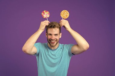 Kafasında mor arka planda lolipoplar olan komik bir adam. Mor arka planda çubuklardaki şekerlemelerle maço bir gülümseme. Mavi tişörtlü, kıvrımlı karamelli manken. Yemek, atıştırmalık ve tatlı yemek.