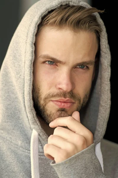 Guy bearded and attractive touches bristle on chin. Masculinity concept. Man with beard or unshaven guy looks handsome hooded. Man with bristle on serious face, urban background, defocused