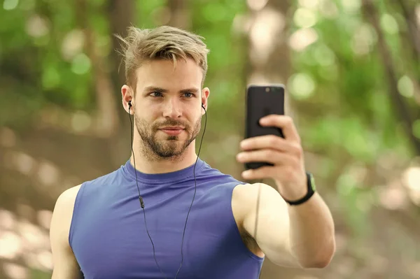 Concetto di gadget sportivo. Atleta cellulare impostare playlist prima di runnig. Atleta uomo volto occupato impostazione app smartphone, sfondo della natura. Allenamento sportivo con pedometro e auricolari — Foto Stock