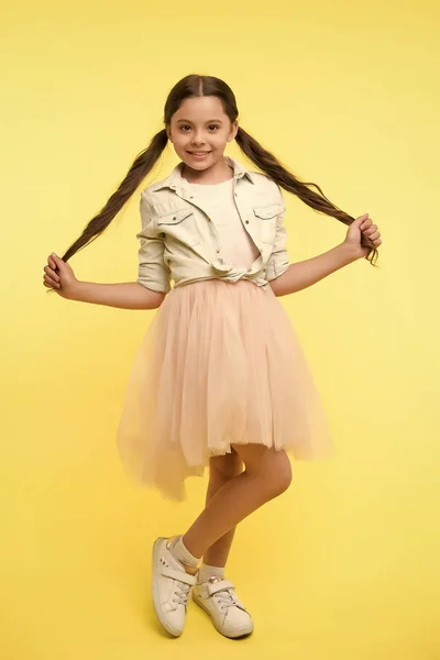 Check out my style. Kid girl charming ponytail hairstyle cute happy yellow background. Child fashionable outfit skirt and denim jacket. Kid stylish fashionable posing holds her long curly hair