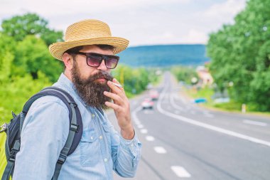 Sigara alışkanlığı. Uzun bir yolculuk önce sigara içilmeyen. Sakal ve bıyık hasır şapka Sigara İçilmeyen, defocused yol arka plan içinde olan adam. Gezgin şık hipster al tut sigara ile