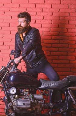 Yenilikçi, vahşi Motorcu deri ceket ciddi yüzüne üzerinde motosiklet üzerinde alır. Erkeksi tutku kavramı. Sakallı, motorcu motorlu bisiklet garaj, tuğla duvar arka plan yakınındaki deri ceketli adam