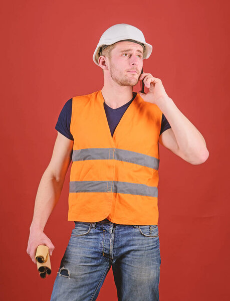 Architect calling engineer to fix plan. Communication concept. Man, foreman in helmet speaking on phone, red background. Engineer, architect, on busy face speaks on smartphone while holds blueprints