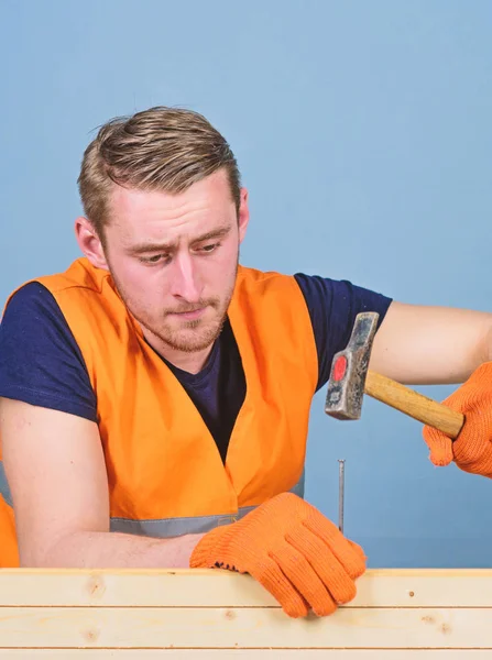 Carpenter concept. Carpenter, woodworker, builder on concentrated face hammering nail into wooden board. Man, handyman in working uniform and protective gloves handcrafting, light blue background
