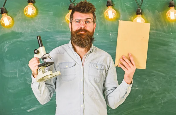 Profesor en anteojos sostiene libro y microscopio. Hombre con barba y bigote en cara estricta en clase. Científico sostiene libro y microscopio, pizarra en el fondo, espacio de copia. Concepto científico — Foto de Stock