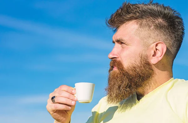 Man with long beard enjoy coffee. Coffee gourmet concept. Man with beard and mustache on strict face drinks coffee, blue sky background, defocused. Bearded man with espresso mug, drinks coffee