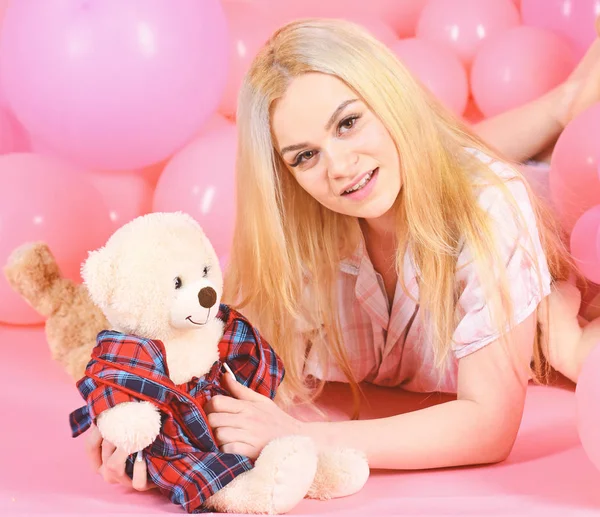 Blonde on smiling face relaxing with teddy bear toy. Woman cute celebrate birthday with balloons. Girl in pajama, domestic clothes lay near air balloons, pink background. Birthday girl concept