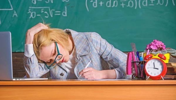 Sigo trabajando. Trabajar mucho más allá del día escolar actual. La cara cansada del maestro sigue trabajando después de clases. Profesor ocupado con el papeleo y la investigación. Profesor mujer sentarse mesa aula pizarra fondo — Foto de Stock