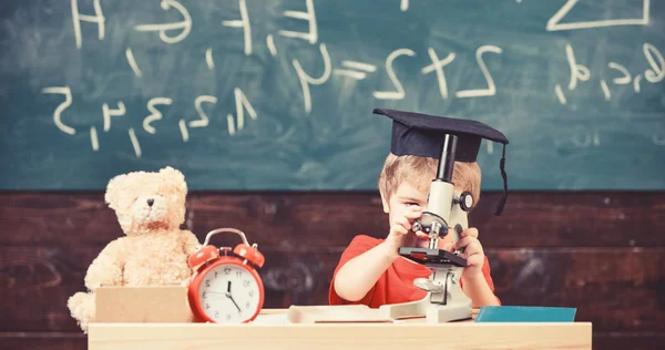 Kid boy i akademiska cap arbete med mikroskopet i klassrummet, svarta tavlan på bakgrund. Barn på upptagen ansikte nära mikroskopet. Smart unge koncept. Första f.d. intresserad av att studera, lärande, utbildning — Stockfoto