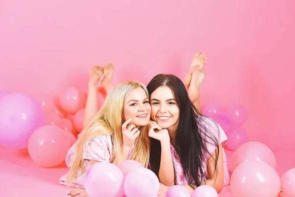 Rubia y morena en caras sonrientes se divierten en la fiesta doméstica. Las niñas yacían sobre el vientre cerca de globos, fondo rosa. Concepto de amistad. Hermanas o mejores amigos en pijama en fiesta de pijamas de niña —  Fotos de Stock