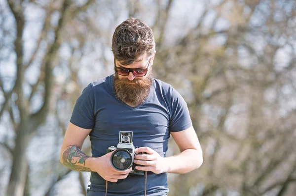 Bearded man works with vintage camera. Man with beard and mustache on concentrated face, branches on background, defocused. Photographer concept. Man with long beard busy with shooting photos — Stock Photo, Image