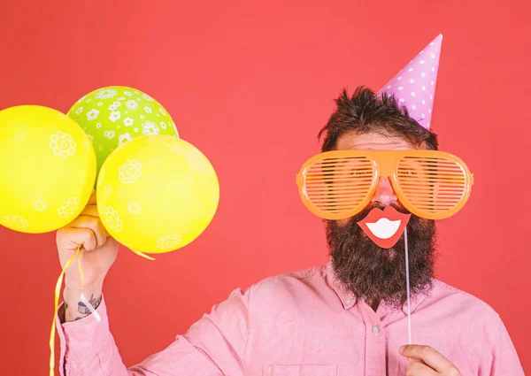 Hipster i giant solglasögon firar. Killen i partiet hatt med air ballonger firar. Foto monter kul koncept. Mannen med skägg på glada ansikte innehar leende mun på pinne, röd bakgrund — Stockfoto