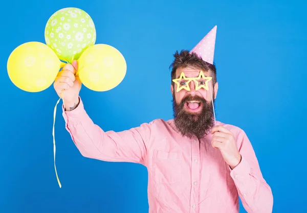 Мужчина с длинной бородой в праздничной кепке веселится с яркими шариками. Арт-перфомер развлекает гостей на детском мероприятии. Бородатый мужчина с бумажными аксессуарами для вечеринки в розовой рубашке на синем фоне — стоковое фото