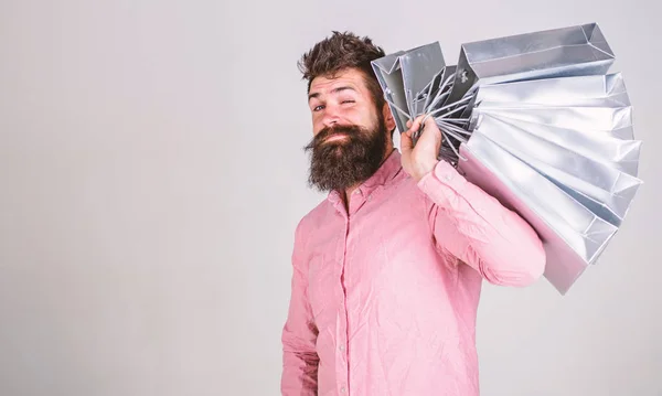 Hipster en guiñar la cara de compras. Concepto de compras. Chico de compras en temporada de ventas, lleva un montón de bolsas en el hombro. Hombre con barba y bigote sostiene bolsas de compras, fondo gris —  Fotos de Stock